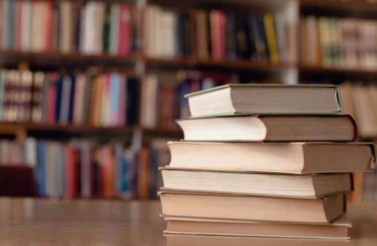 A pile of books on a table with filled bookshelves behind out of focus
