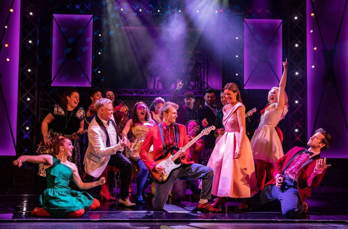 The cast of Footloose gathered on stage watching a man serenade a woman with an electric guitar