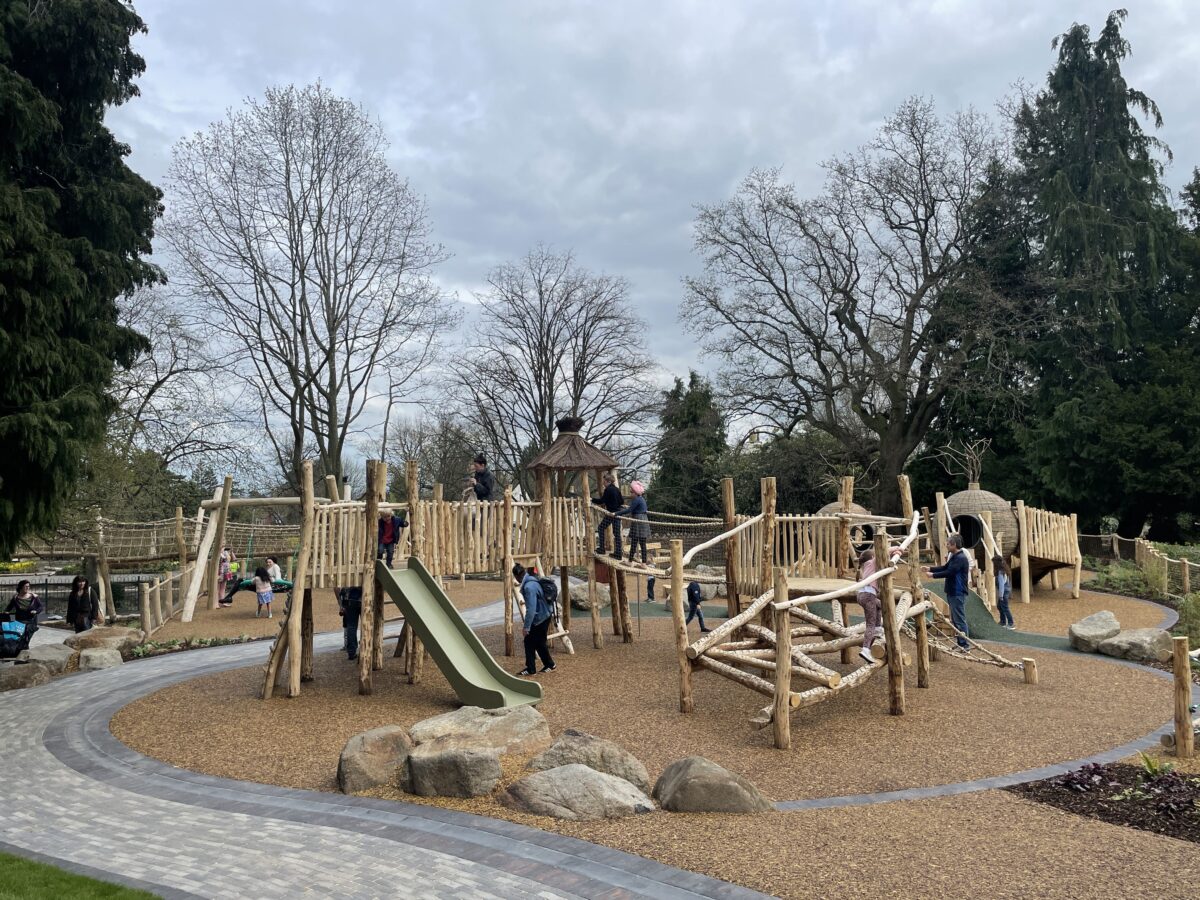 Priory Park Play Area overview showing families playing on the swing, slides and wooden climbing apparatus