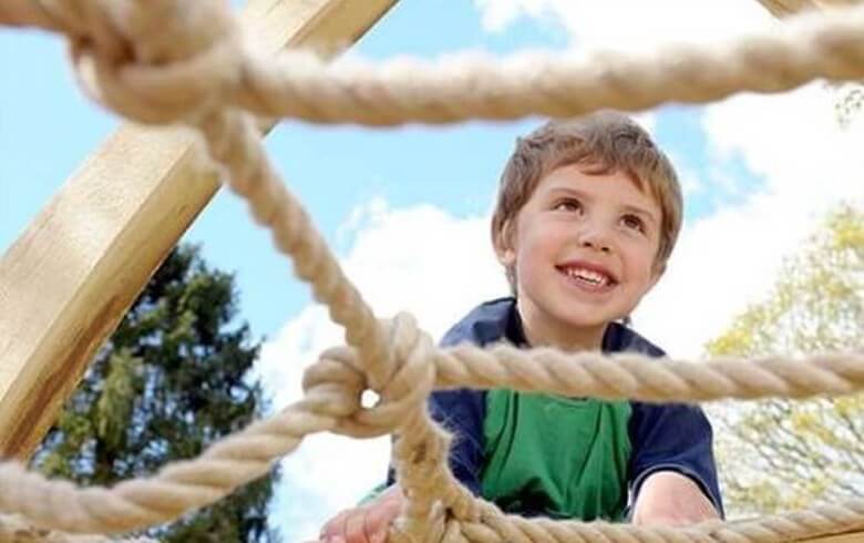 A child smiles and climbs on a rope frame at Witley Court Play Area
