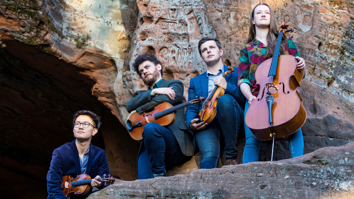 4 musicians holding 3 violins and 1 cello stood on a rock