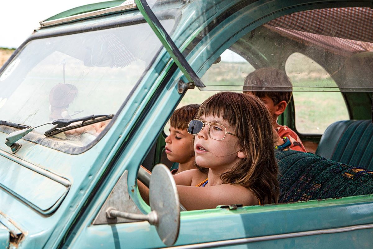 Three Children sit in a car, one is driving. The driver has shaggy brown hair and is wearing sunglasses with one lens missing