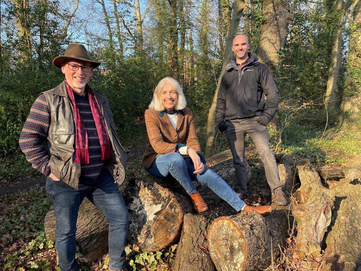 Three people pose in a wooded area upon a pile of logs
