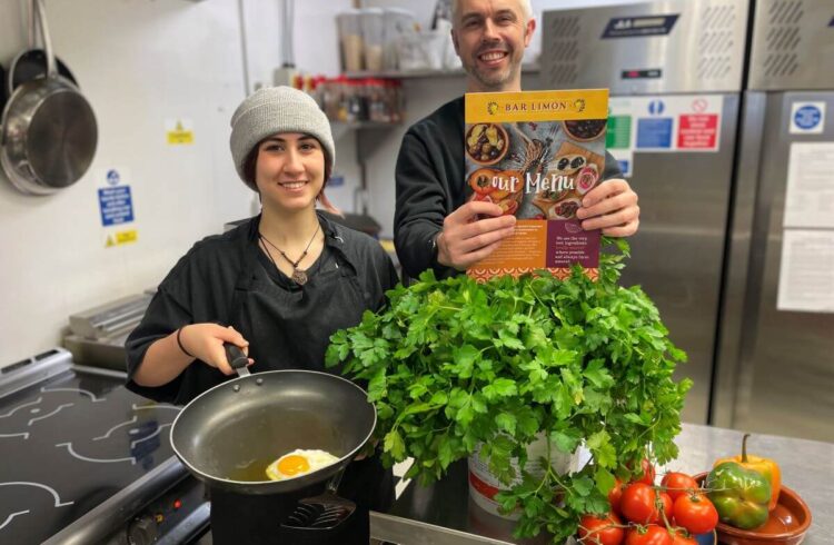 Two people in a commercial kitchen, one holds a frying pan with a fried egg and the other a menu. There is fresh produce around them including coriander and tomatoes.