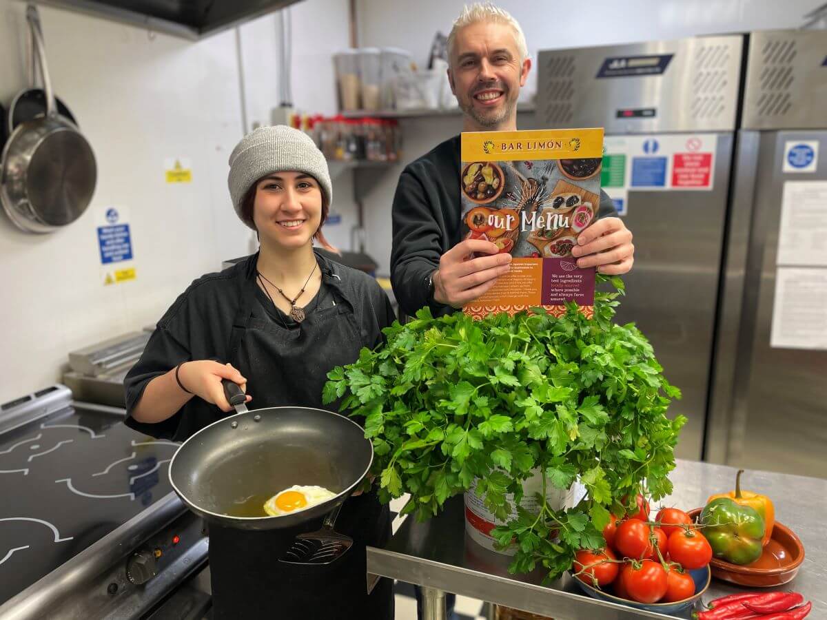 Two people in a commercial kitchen, one holds a frying pan with a fried egg and the other a menu. There is fresh produce around them including coriander and tomatoes.