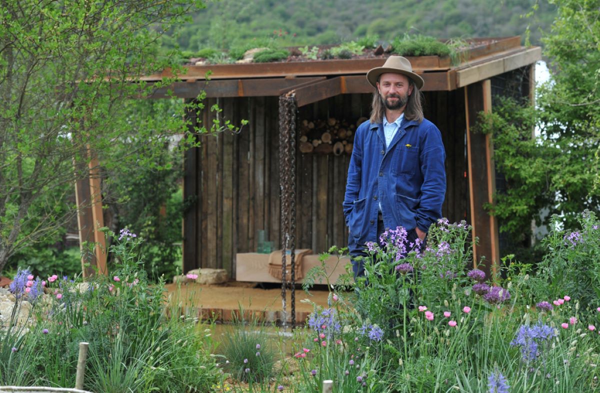 A man in a wide brimmed had an blue workwear stands in a garden