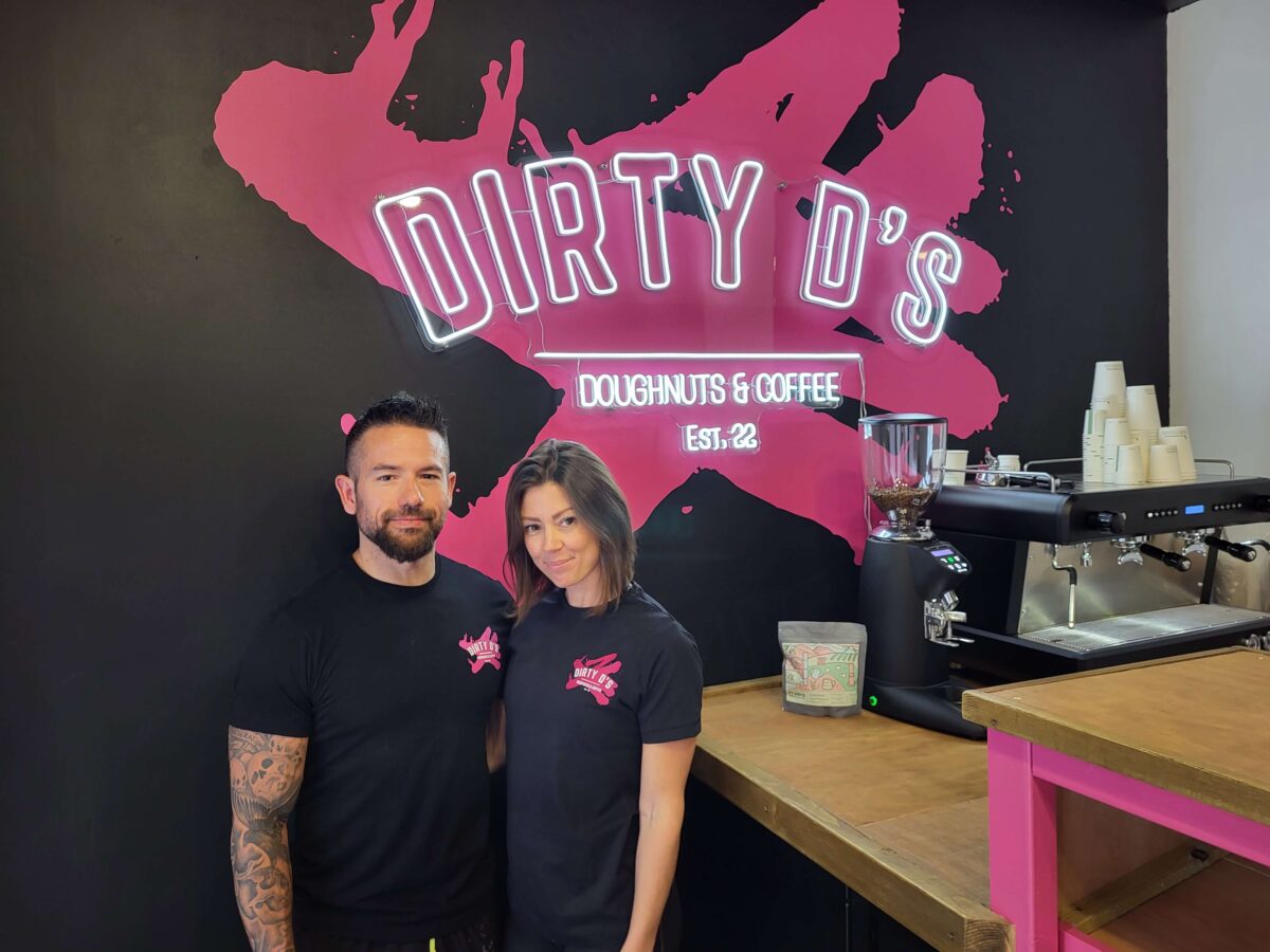 Two people standing in a café with a pink and black logo on the wall behind