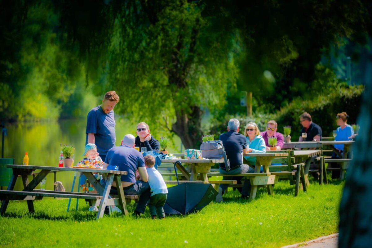 People dine on picnic benches on a green river bank