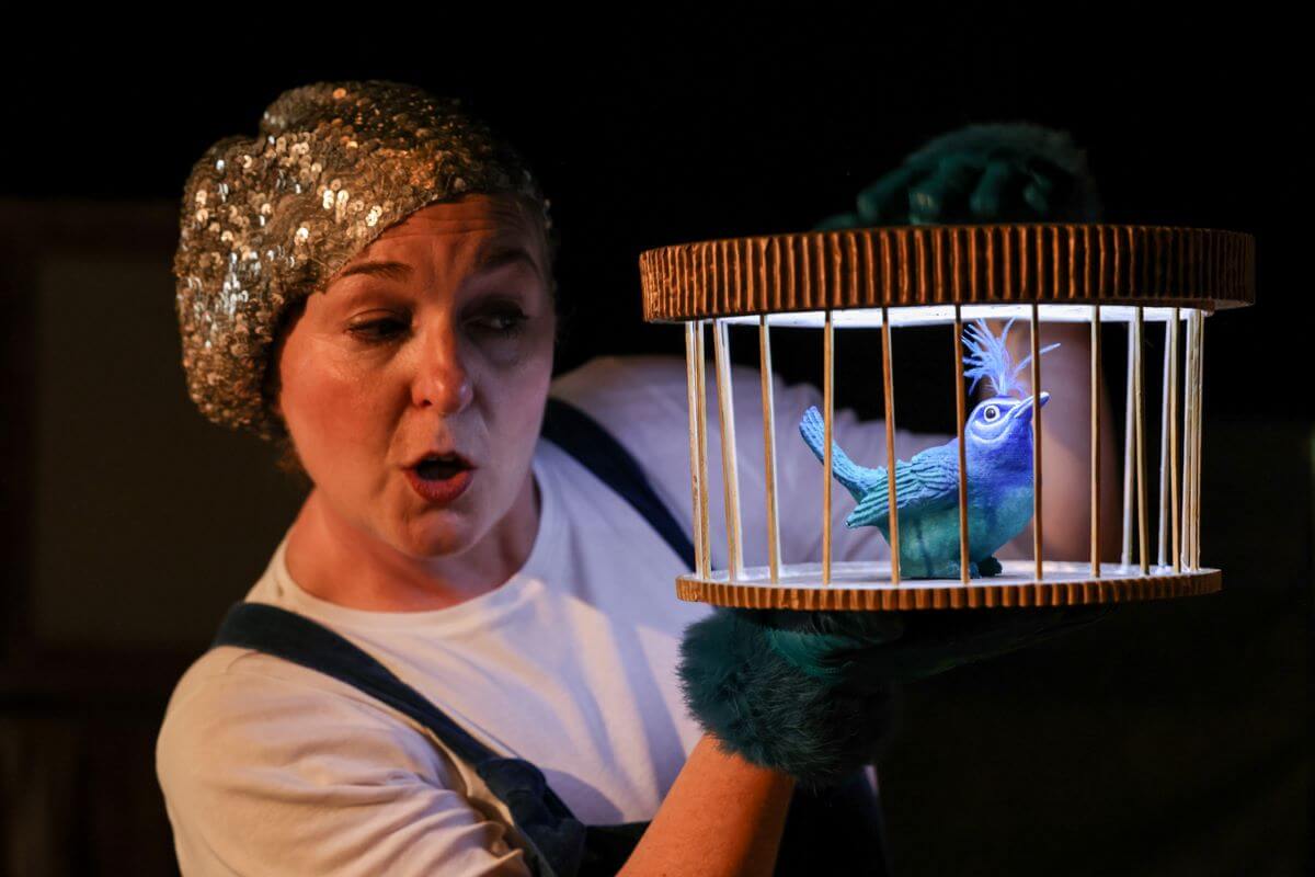 A person in a sequin beret holds a puppet bird in a cage