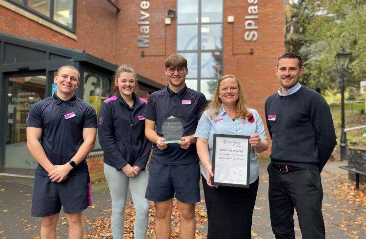 five people stand outside a brick leisure centre holding a certificate and smiling