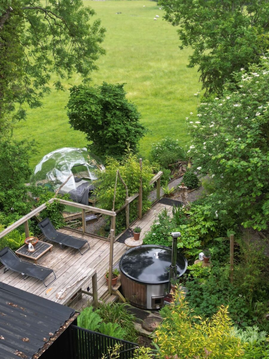An aerial shot of the outdoor area of a cabin surrounded by trees with a hot tub, geodome and sun loungers.