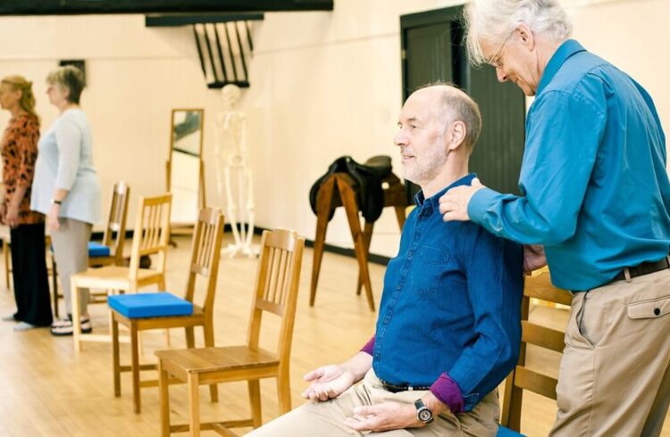 A man in a blue shirt, seated on a chair in a wooden-floored room, being shown Alexander Technique