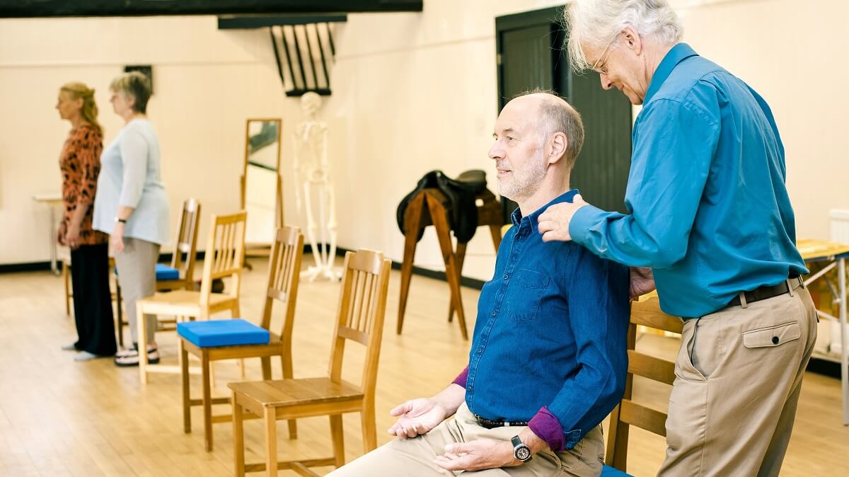 A man in a blue shirt, seated on a chair in a wooden-floored room, being shown Alexander Technique