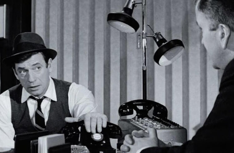 A still from the film showing two men seated near a lamp and telephones