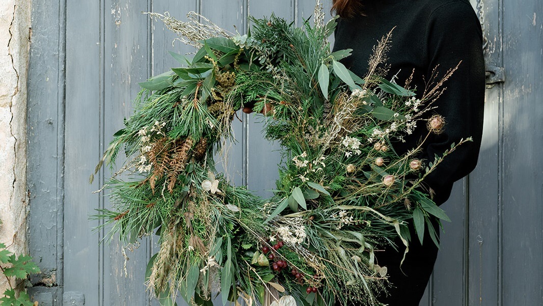 A Christmas wreath made from natural materials being held in front of a green wooden door
