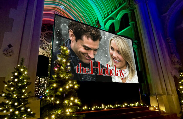 A big screen in an ornate chapel with Christmas trees, showing an advert for the film ' The Holiday'