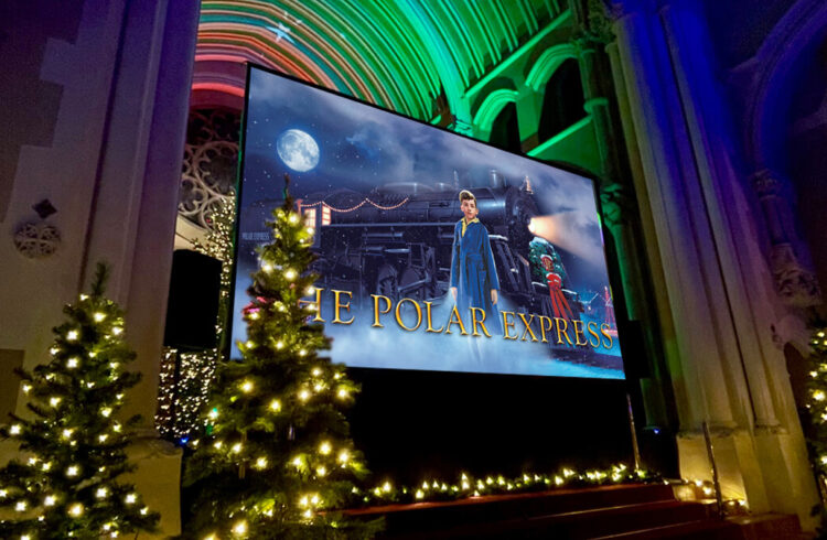 A big screen in an ornate chapel with Christmas trees, showing an advert for the film 'The Polar Express'