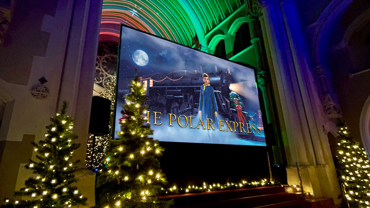 A big screen in an ornate chapel with Christmas trees, showing an advert for the film 'The Polar Express'