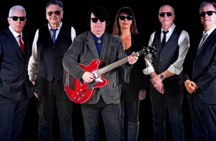 Six smartly dressed musicians (five male and one female), all wearing sunglasses and one holding a guitar