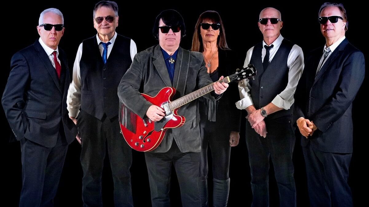 Six smartly dressed musicians (five male and one female), all wearing sunglasses and one holding a guitar